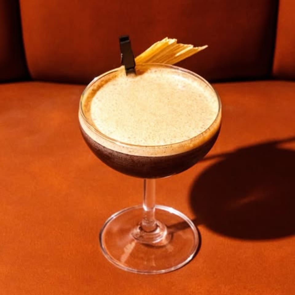 A dark cocktail with a foamy top, garnished with dried pasta, sits on an orange surface.