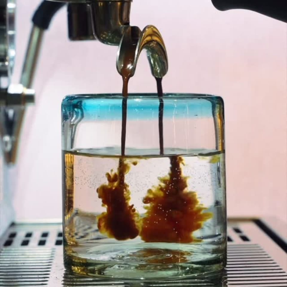 Espresso pours from a machine into a glass of water, creating beautiful brown swirls.