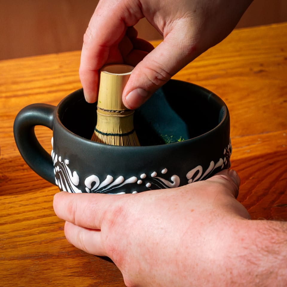 Hands whisking matcha in a black ceramic mug with white designs on a wooden table.